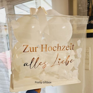 May include: A clear, square gift box filled with white balloons. The box has a white bow and the words "Zur Hochzeit alles Liebe" in gold lettering. The box is decorated with white heart-shaped cutouts.