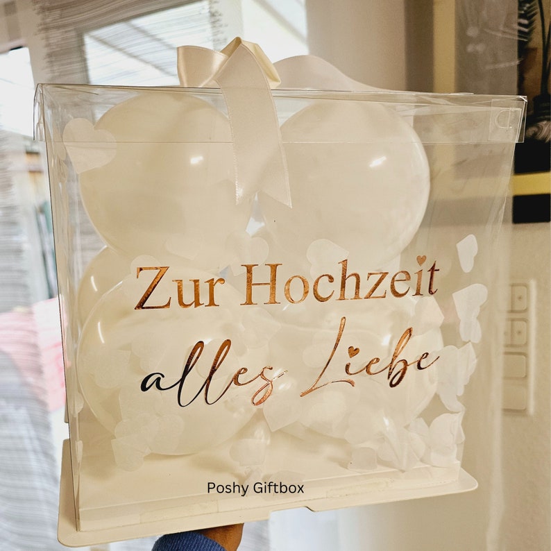 May include: A clear, square gift box filled with white balloons. The box has a white bow and the words "Zur Hochzeit alles Liebe" in gold lettering. The box is decorated with white heart-shaped cutouts.