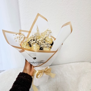 May include: A bouquet of golden chocolate truffles wrapped in white and gold paper with a gold ribbon. The bouquet is decorated with dried white flowers and a gold glitter leaf. The bouquet is held by a hand with a brown sweater.