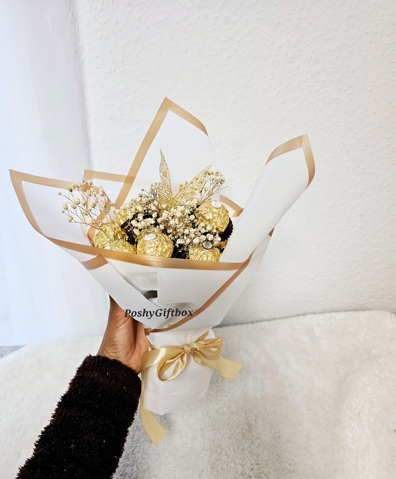 May include: A bouquet of golden chocolate truffles wrapped in white and gold paper with a gold ribbon. The bouquet is decorated with dried white flowers and a gold glitter leaf. The bouquet is held by a hand with a brown sweater.