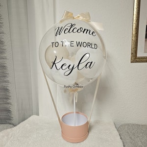 May include: A clear balloon hot air balloon with white and cream balloons inside. The balloon has the text "Welcome to the world Keyla" printed on it. The balloon is attached to a pink box with a white ribbon.