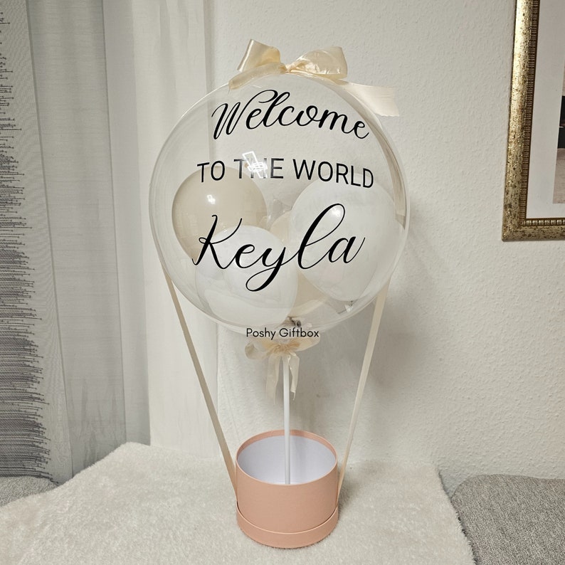May include: A clear balloon hot air balloon with white and cream balloons inside. The balloon has the text "Welcome to the world Keyla" printed on it. The balloon is attached to a pink box with a white ribbon.