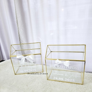 May include: Two clear glass boxes with gold frames and white ribbons. The boxes are labeled "Hochzeitspost" with the names and dates of the weddings. The boxes are for wedding cards and gifts.