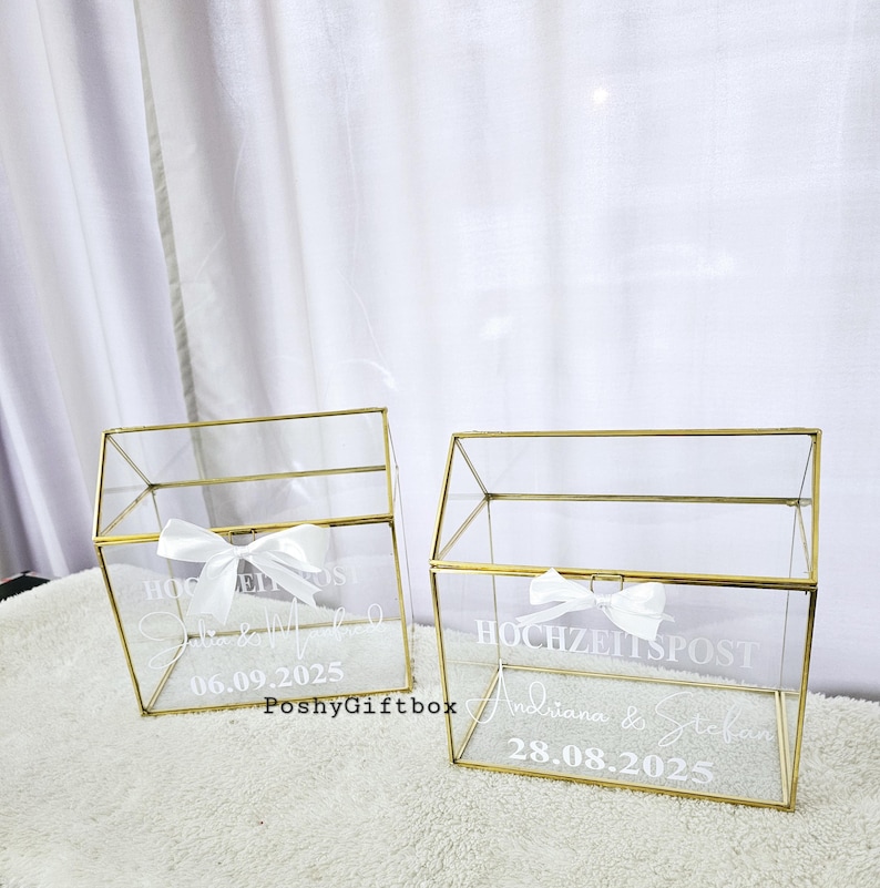 May include: Two clear glass boxes with gold frames and white ribbons. The boxes are labeled "Hochzeitspost" with the names and dates of the weddings. The boxes are for wedding cards and gifts.
