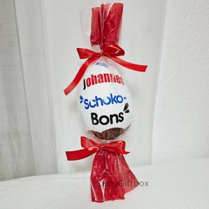Peut inclure: Un &oelig;uf en chocolat blanc envelopp&eacute; de cellophane rouge avec un ruban rouge. L'&oelig;uf est d&eacute;cor&eacute; du texte "Johannes Schoko-Bons".