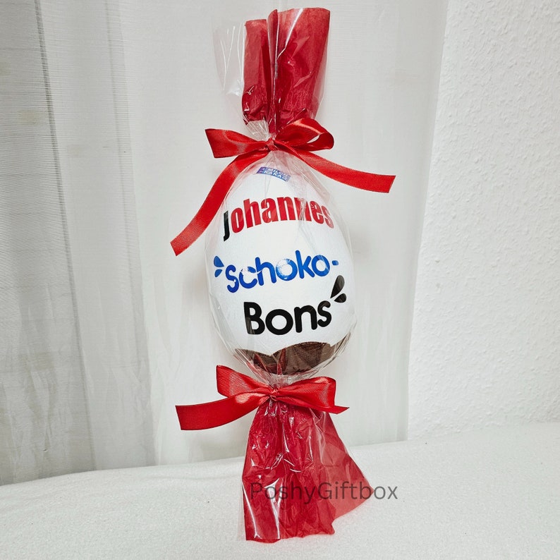 Peut inclure: Un &oelig;uf en chocolat blanc envelopp&eacute; de cellophane rouge avec un ruban rouge. L'&oelig;uf est d&eacute;cor&eacute; du texte "Johannes Schoko-Bons".