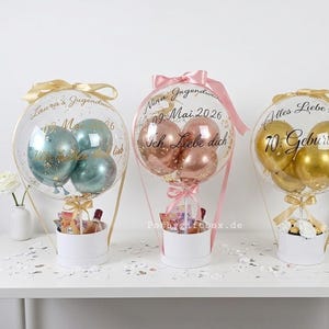 Op de afbeelding: Drie transparante ballongeschenken in witte cilindrische dozen. De linkerballon bevat blauwgroene ballonnen met de tekst "Laura's Jugendweihe 09. Mai 2026". Het midden bevat ros&eacute;gouden ballonnen met "Nina Jugendweihe 09. Mai 2026 Ich Liebe dich". De rechter bevat gouden ballonnen met "Alles Liebe zum 70. Geburtstag".