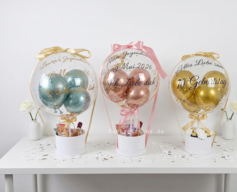 Op de afbeelding: Drie transparante ballongeschenken in witte cilindrische dozen. De linkerballon bevat blauwgroene ballonnen met de tekst "Laura's Jugendweihe 09. Mai 2026". Het midden bevat ros&eacute;gouden ballonnen met "Nina Jugendweihe 09. Mai 2026 Ich Liebe dich". De rechter bevat gouden ballonnen met "Alles Liebe zum 70. Geburtstag".