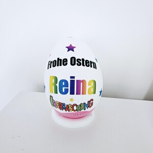 Op de afbeelding: Een wit paasei met de tekst "Frohe Ostern" en de naam "Reina" in regenboogkleuren. Het ei heeft ook een ster en de tekst "&Uuml;berraschung" in kleurrijke letters. Het ei staat op een roze voet met de tekst "PoshyGiftbox".