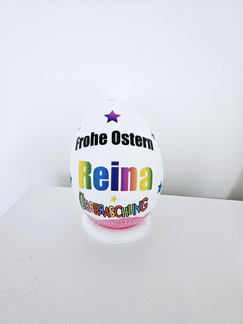 Op de afbeelding: Een wit paasei met de tekst "Frohe Ostern" en de naam "Reina" in regenboogkleuren. Het ei heeft ook een ster en de tekst "&Uuml;berraschung" in kleurrijke letters. Het ei staat op een roze voet met de tekst "PoshyGiftbox".