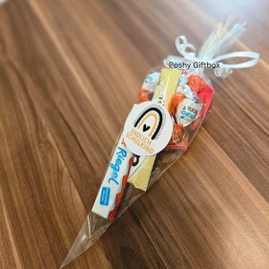 May include: A clear plastic cone filled with candy and a chocolate bar. The cone is tied with a white ribbon and has a sticker that says "Poshy Giftbox" and another sticker that says "Endlich Schulkind" with a rainbow and heart design.