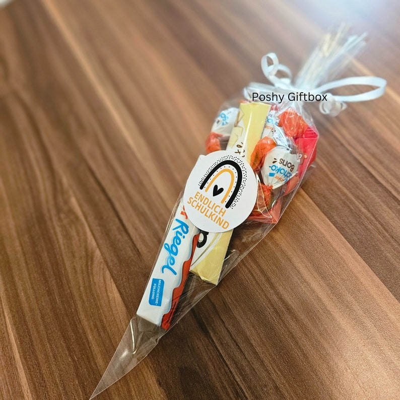 May include: A clear plastic cone filled with candy and a chocolate bar. The cone is tied with a white ribbon and has a sticker that says "Poshy Giftbox" and another sticker that says "Endlich Schulkind" with a rainbow and heart design.