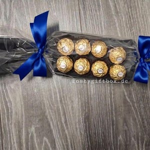 May include: A clear cellophane gift cracker containing eight round chocolates wrapped in gold foil. The packaging is secured at both ends with vibrant royal blue satin ribbons. The text poshygiftbox.de is printed in white on the clear plastic.