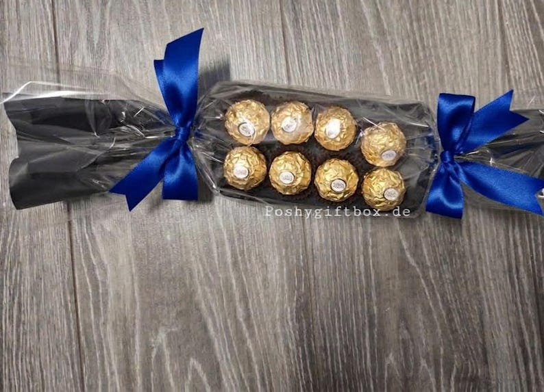 May include: A clear cellophane gift cracker containing eight round chocolates wrapped in gold foil. The packaging is secured at both ends with vibrant royal blue satin ribbons. The text poshygiftbox.de is printed in white on the clear plastic.