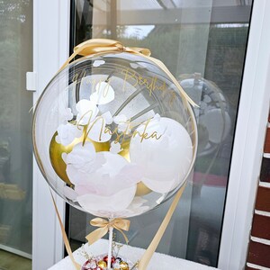 May include: A clear balloon with gold lettering that says "Happy Birthday Nastinka" and white and gold balloons inside. The balloon is attached to a white heart-shaped box filled with chocolates and tied with a gold ribbon. The box is labeled "PoshyGiftbox".