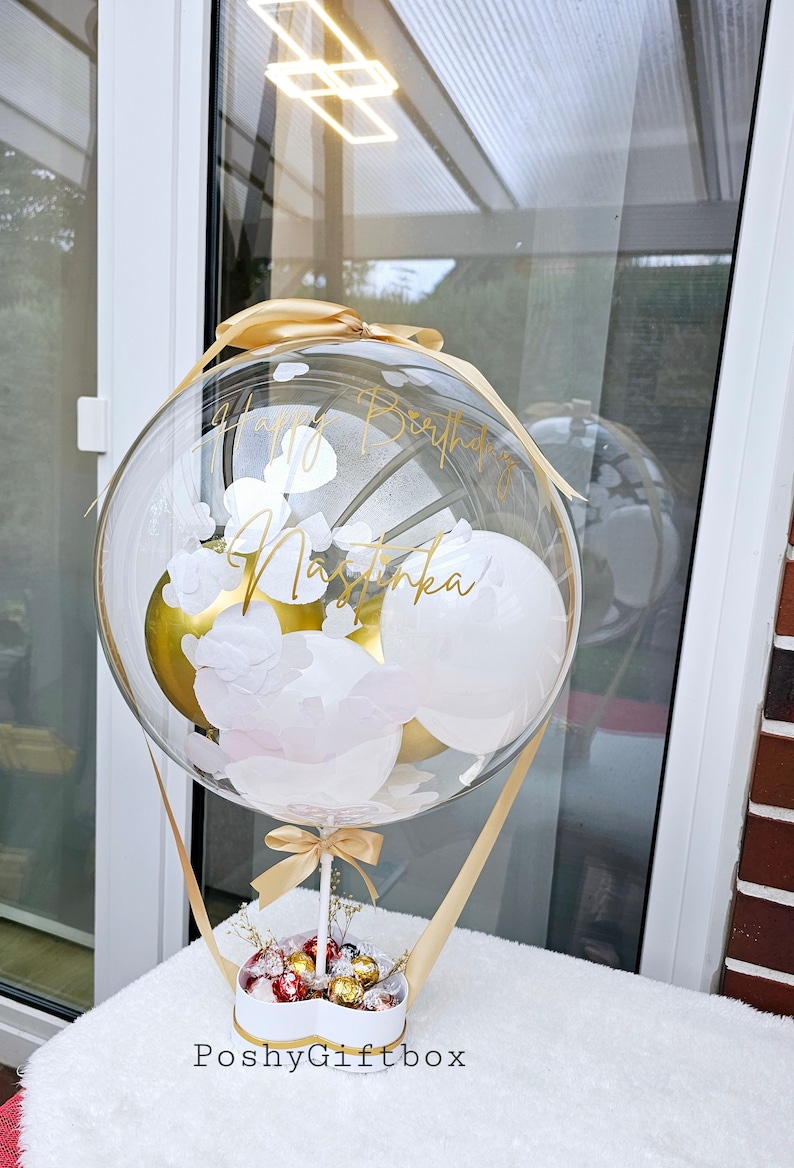 May include: A clear balloon with gold lettering that says "Happy Birthday Nastinka" and white and gold balloons inside. The balloon is attached to a white heart-shaped box filled with chocolates and tied with a gold ribbon. The box is labeled "PoshyGiftbox".