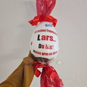 Peut inclure: Un &oelig;uf en chocolat blanc envelopp&eacute; de cellophane rouge avec un ruban rouge. L'&oelig;uf est d&eacute;cor&eacute; du texte "Pour mon homme de r&ecirc;ve Lars. Tu es mon grand amour."
