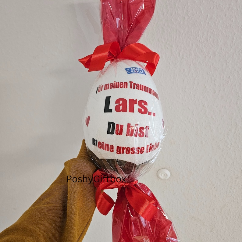 Peut inclure: Un &oelig;uf en chocolat blanc envelopp&eacute; de cellophane rouge avec un ruban rouge. L'&oelig;uf est d&eacute;cor&eacute; du texte "Pour mon homme de r&ecirc;ve Lars. Tu es mon grand amour."