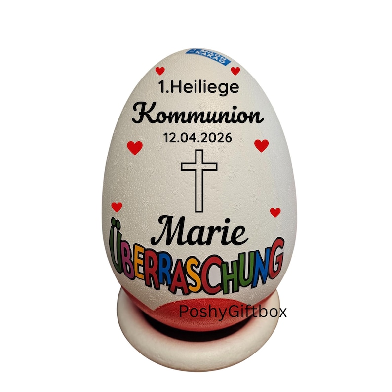 May include: A white egg-shaped decorative item with text and a cross. The text includes "1. Heilige Kommunion", "12.04.2026", and "Marie &Uuml;berraschung". Red heart accents are scattered around the egg.