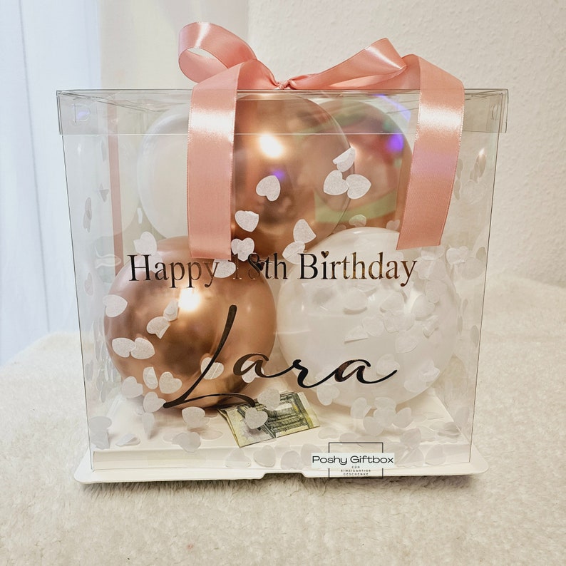 May include: A clear gift box with a pink ribbon and balloons. The balloons are white, rose gold, and iridescent. The box has the text "Happy 8th Birthday Lara" and heart-shaped confetti. The bottom of the box has a small gift.
