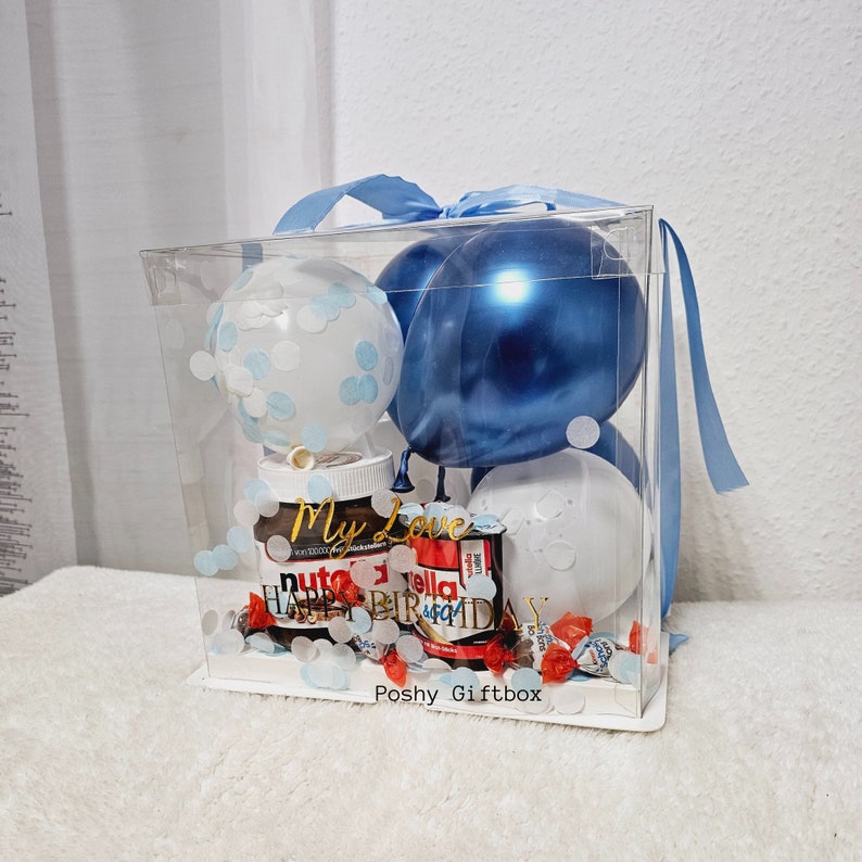May include: A clear plastic gift box with a blue ribbon tied around it. Inside the box are three balloons, two blue and one white with blue polka dots. There is a jar of Nutella and a chocolate bar with the words "Happy Birthday" on it. The box also contains confetti and candy.
