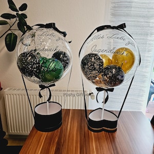 May include: Two clear balloon hot air balloons with black ribbons and black and gold confetti balloons inside. The balloons are attached to black boxes with white lids. The left balloon has the text "Alles Liebe zur Jugendweihe" and the right balloon has the text "Alles Liebe zur Jugendweihe".