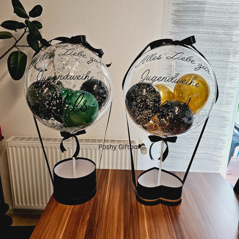 May include: Two clear balloon hot air balloons with black ribbons and black and gold confetti balloons inside. The balloons are attached to black boxes with white lids. The left balloon has the text "Alles Liebe zur Jugendweihe" and the right balloon has the text "Alles Liebe zur Jugendweihe".