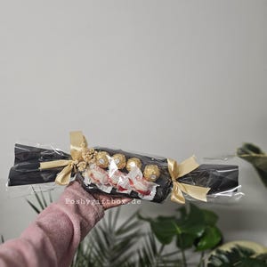 May include: A gift-wrapped sweet-shaped package featuring black paper, clear cellophane, and gold ribbon bows at each end. Inside are four Raffaello coconut almond confections. The text Poshygiftbox.de is printed on the clear plastic wrapping.