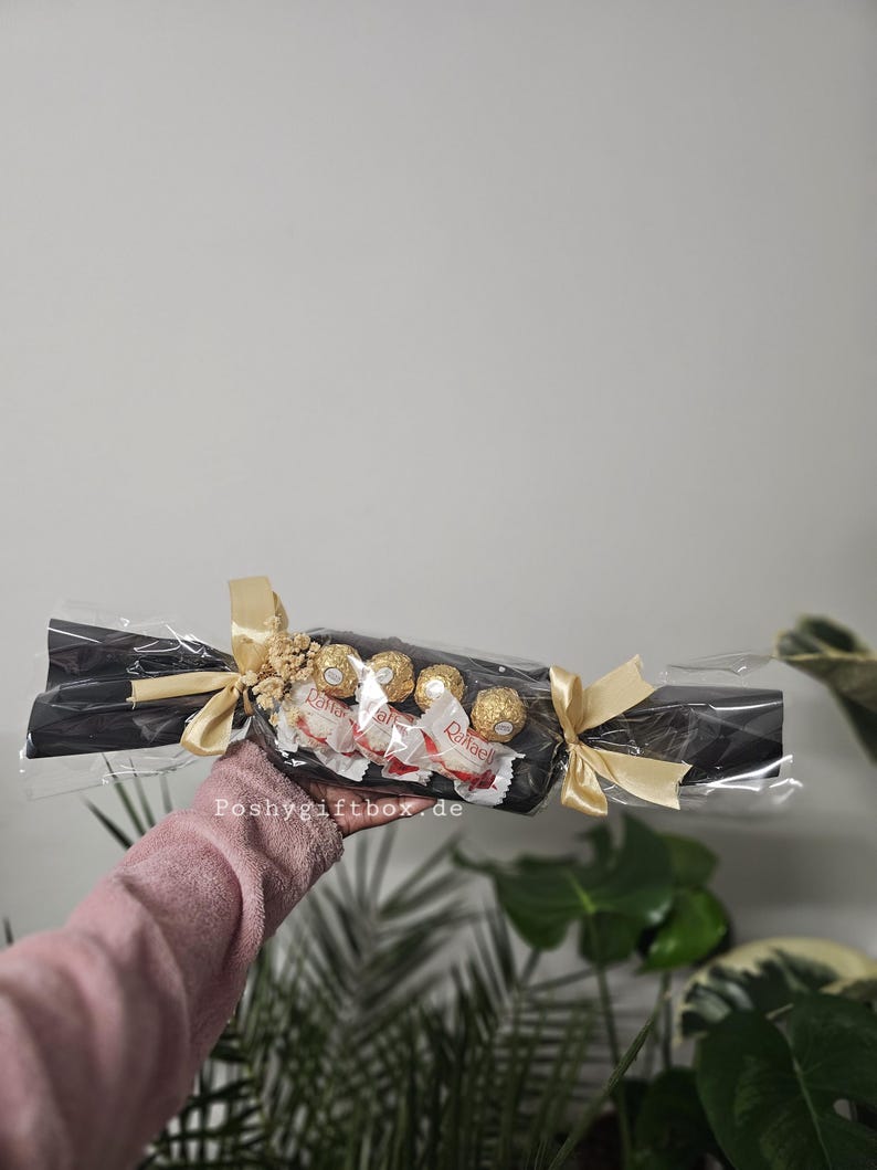 May include: A gift-wrapped sweet-shaped package featuring black paper, clear cellophane, and gold ribbon bows at each end. Inside are four Raffaello coconut almond confections. The text Poshygiftbox.de is printed on the clear plastic wrapping.