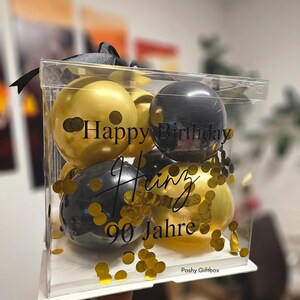 May include: A clear box filled with black and gold balloons with gold confetti. The balloons have the text "Happy Birthday Heinz 90 Jahre" printed on them.