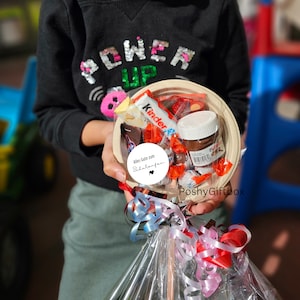 May include: A gift basket filled with various chocolate candies, a jar of Nutella, and a Kinder chocolate bar. The basket is wrapped in clear cellophane and tied with a red and blue ribbon. A white sticker with the text "Alles Gute zum Schulanfang" is attached to the basket.