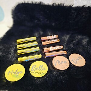 Pu&ograve; includere: Una collezione di tubetti di rossetto e specchietti compatti personalizzati in oro e oro rosa con nomi come Bella, Helga, Doris e Nina. Gli articoli sono disposti su una superficie di pelliccia sintetica nera.