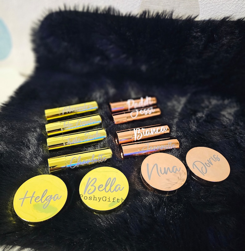 Pu&ograve; includere: Una collezione di tubetti di rossetto e specchietti compatti personalizzati in oro e oro rosa con nomi come Bella, Helga, Doris e Nina. Gli articoli sono disposti su una superficie di pelliccia sintetica nera.