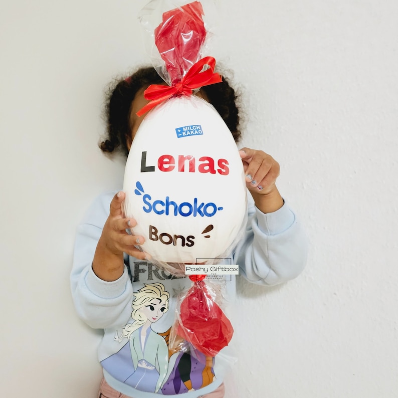 Peut inclure: Un &oelig;uf en chocolat blanc avec des accents rouges et bleus. L'&oelig;uf est envelopp&eacute; dans du plastique transparent et a un ruban rouge. L'&oelig;uf est d&eacute;cor&eacute; du texte "Lenas Schoko-Bons" et "Milch Kakao".