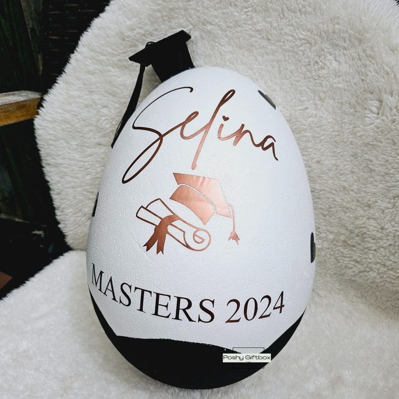 May include: White egg-shaped object with a black graduation cap on top. The egg is decorated with a gold graduation cap, diploma, and the name "Selina" in cursive. The text "Masters 2024" is written in black below the design.