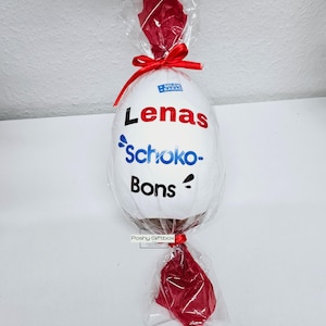 Peut inclure: Un &oelig;uf en chocolat blanc envelopp&eacute; de cellophane rouge avec un ruban rouge. L'&oelig;uf est d&eacute;cor&eacute; d'un autocollant qui dit "Lenas Schoko-Bons".