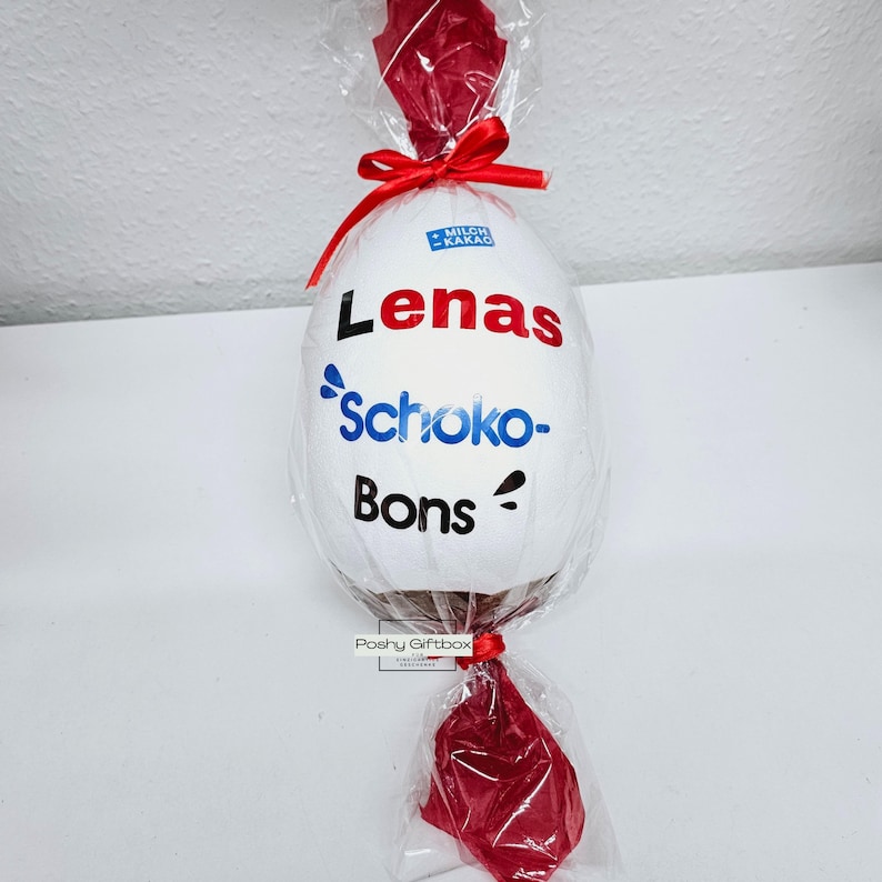 Peut inclure: Un &oelig;uf en chocolat blanc envelopp&eacute; de cellophane rouge avec un ruban rouge. L'&oelig;uf est d&eacute;cor&eacute; d'un autocollant qui dit "Lenas Schoko-Bons".