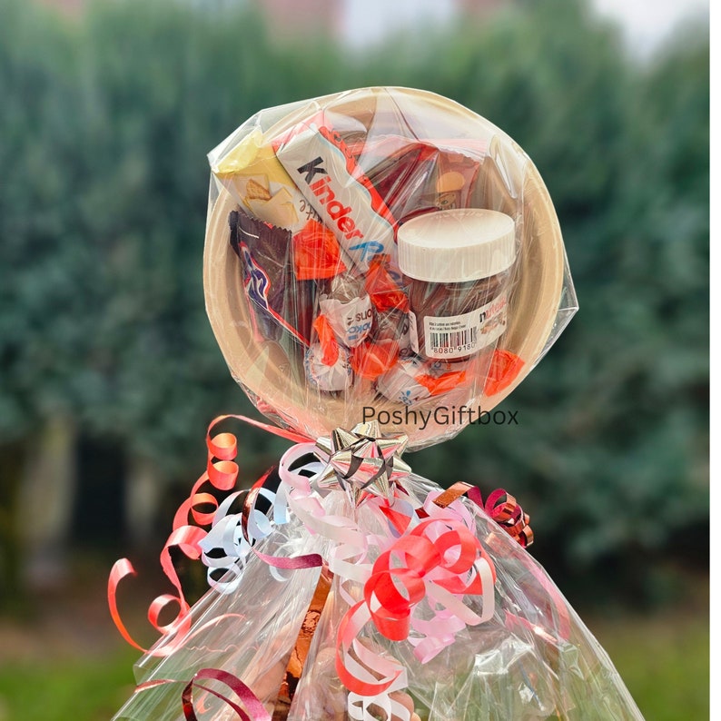 May include: A clear plastic container filled with assorted chocolate sweets, a small jar of Nutella, and a Kinder chocolate bar. The container is wrapped in clear cellophane and tied with red, pink and white ribbons. The text "PoshyGiftbox" is visible on the container.