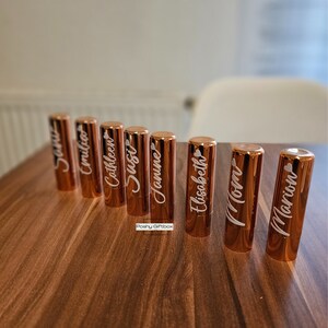 Pu&ograve; includere: Otto tubetti di rossetto in oro rosa con nomi personalizzati, tra cui "Susie", "Jamie", "Mamma" e "Marion", su una superficie in legno.