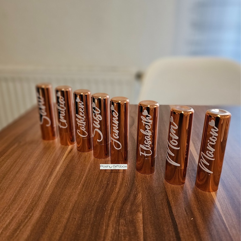 Pu&ograve; includere: Otto tubetti di rossetto in oro rosa con nomi personalizzati, tra cui "Susie", "Jamie", "Mamma" e "Marion", su una superficie in legno.