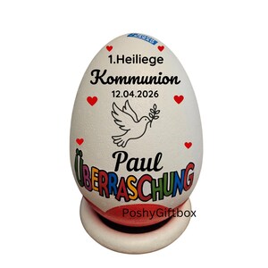 May include: A white, egg-shaped decorative item with text and graphics. The text includes "1. Heilige Kommunion", the date "12.04.2026", and the name "Paul". A dove graphic is also present, along with red heart accents.