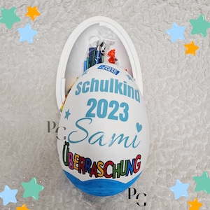 Peut inclure: Un contenant en forme d'&oelig;uf blanc avec un couvercle bleu. Le contenant est d&eacute;cor&eacute; des mots "Schulkind 2023 Sami" et "&Uuml;berraschung".