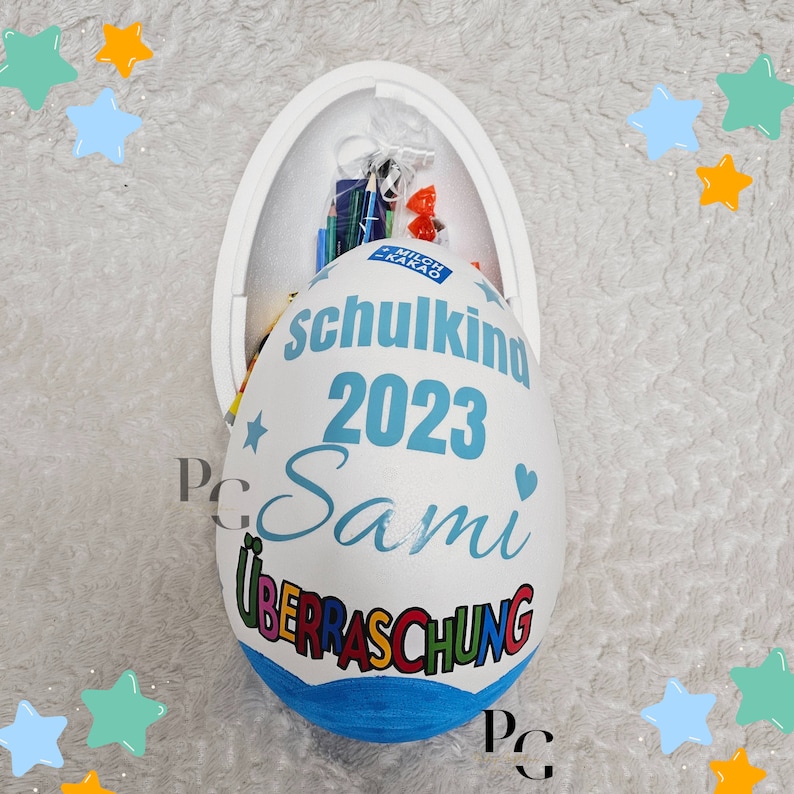 Peut inclure: Un contenant en forme d'&oelig;uf blanc avec un couvercle bleu. Le contenant est d&eacute;cor&eacute; des mots "Schulkind 2023 Sami" et "&Uuml;berraschung".