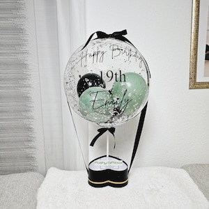 May include: A clear balloon with black and green balloons inside. The balloon has a black ribbon and the text "Happy Birthday 19th Emily" written on it. The balloon is attached to a black heart-shaped box with the text "Poshy Giftbox" written on it.