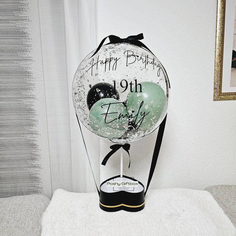 May include: A clear balloon with black and green balloons inside. The balloon has a black ribbon and the text "Happy Birthday 19th Emily" written on it. The balloon is attached to a black heart-shaped box with the text "Poshy Giftbox" written on it.