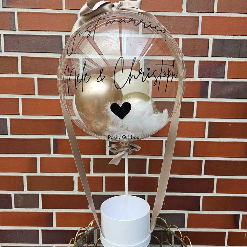 May include: A clear balloon with a white ribbon and a white box at the bottom. The balloon has gold and white balloons inside and a black heart. The balloon has the text "Just married" and "Nile & Christoph" written on it. The box has the text "Poshy Giftbox" written on it.