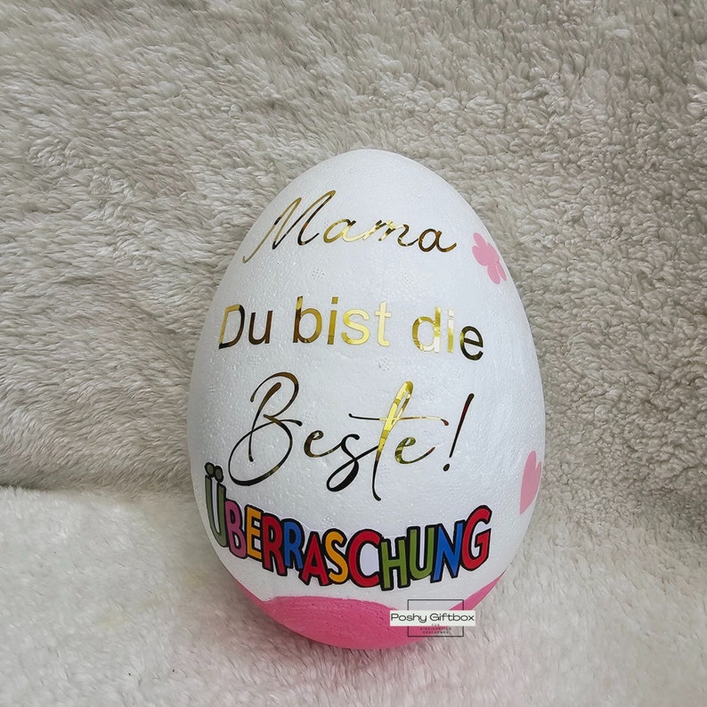May include: White styrofoam egg with gold lettering that reads "Mama Du bist die Beste!" and colorful lettering that reads "Ueberraschung". The egg is decorated with pink hearts and flowers.