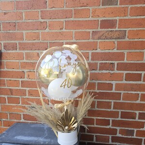May include: A clear balloon filled with smaller gold and white balloons, featuring the gold text "Rahmiye" and "40" on the surface. The balloon is anchored in a white cylindrical base decorated with dried pampas grass and a gold fan-shaped accent.