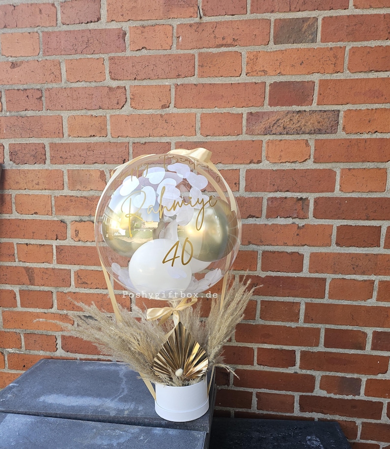 May include: A clear balloon filled with smaller gold and white balloons, featuring the gold text "Rahmiye" and "40" on the surface. The balloon is anchored in a white cylindrical base decorated with dried pampas grass and a gold fan-shaped accent.