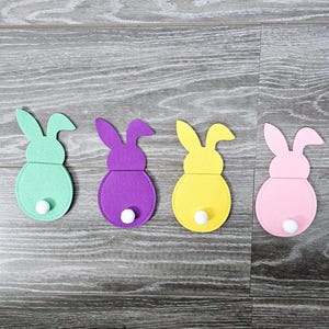 May include: Four felt bunny shapes in different colors: green, purple, yellow, and pink. Each bunny has a white pom-pom tail.
